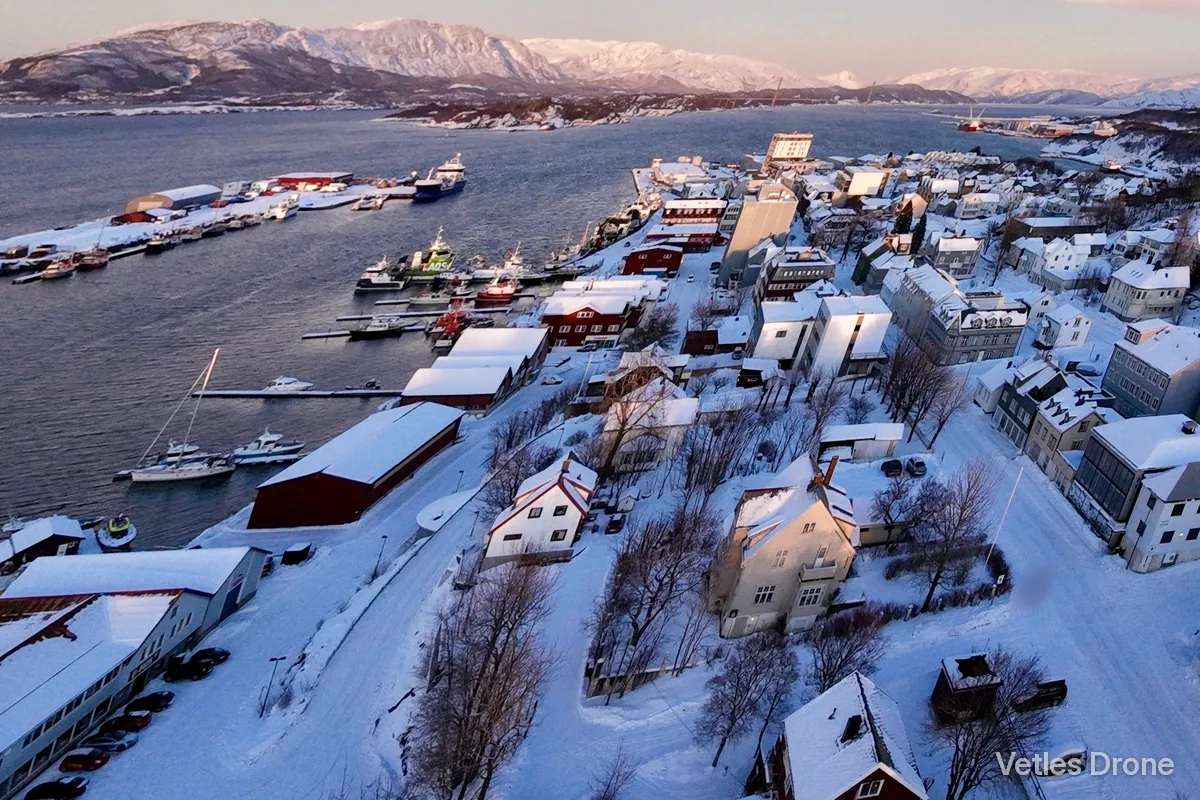Dag: havna og sentrum i Sandnessjøen i vintersol, med snødekte tak, kaier, båter og fjell i bakgrunnen, sett fra drone