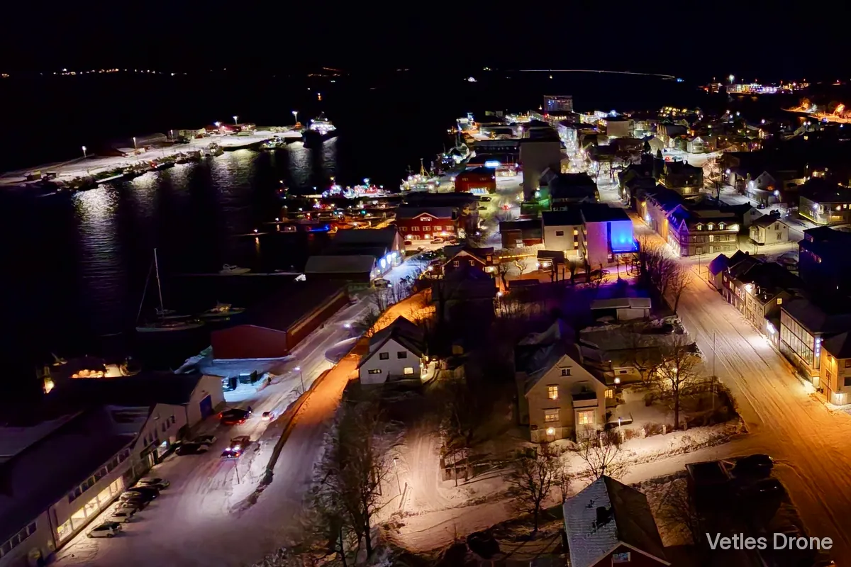 Kveld: havna og sentrum i Sandnessjøen i mørket, med opplyste gater, kaier og båter, sett fra drone
