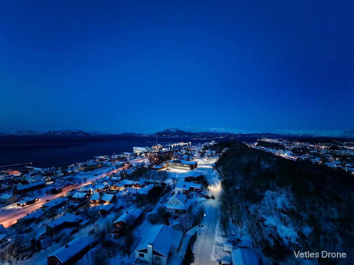 Sandnessjøen i blåtimen en vinterkveld, med lys i gatene, snødekt skog og fjell i horisonten, sett fra drone