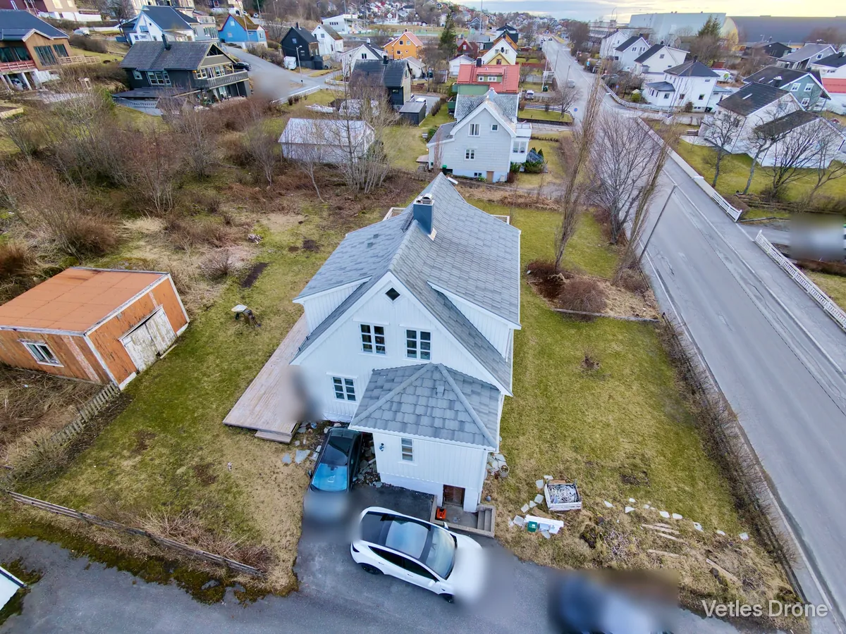 Hvitt hus med grått tak, inngangsparti og hage i et boligområde, fotografert skrått ovenfra med drone