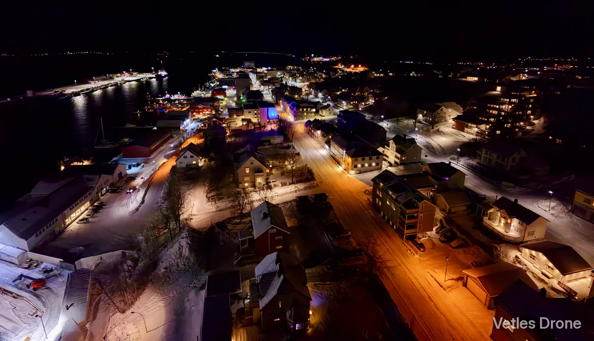 Sandnessjøen en vinterkveld, med opplyste gater, hus og havna i mørket, sett fra drone