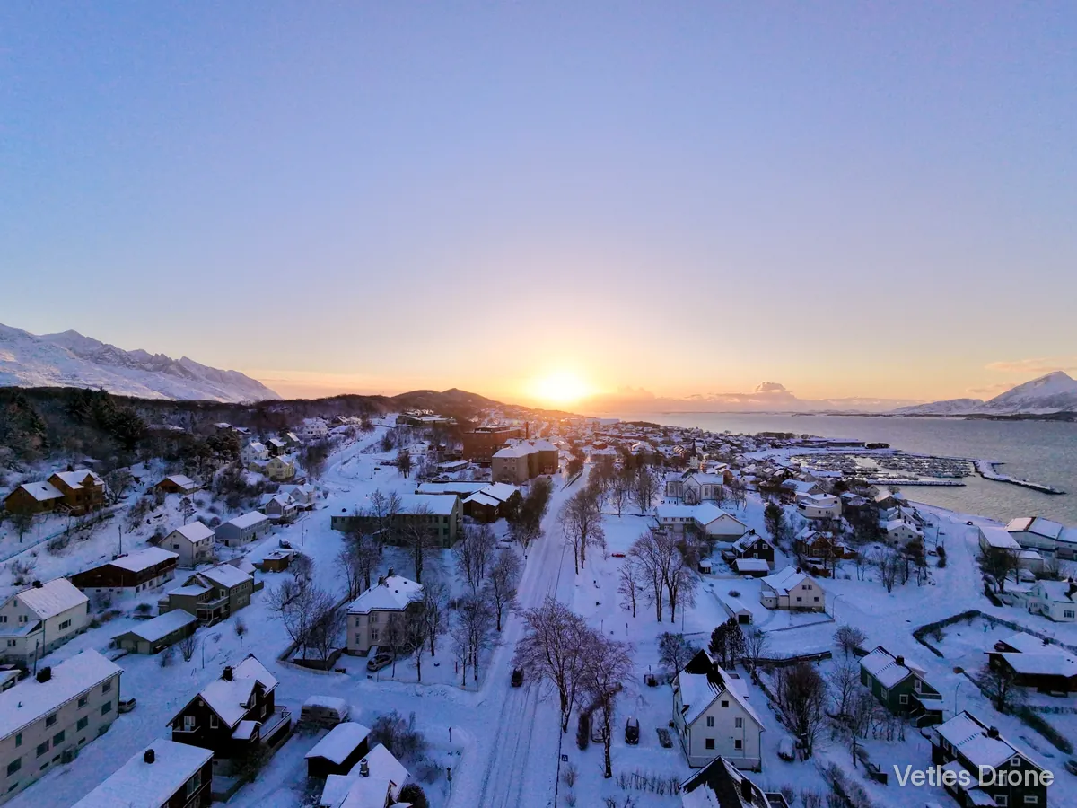 Boligområde i Sandnessjøen dekket av snø i solnedgang, med småbåthavn og fjell i bakgrunnen, sett fra drone