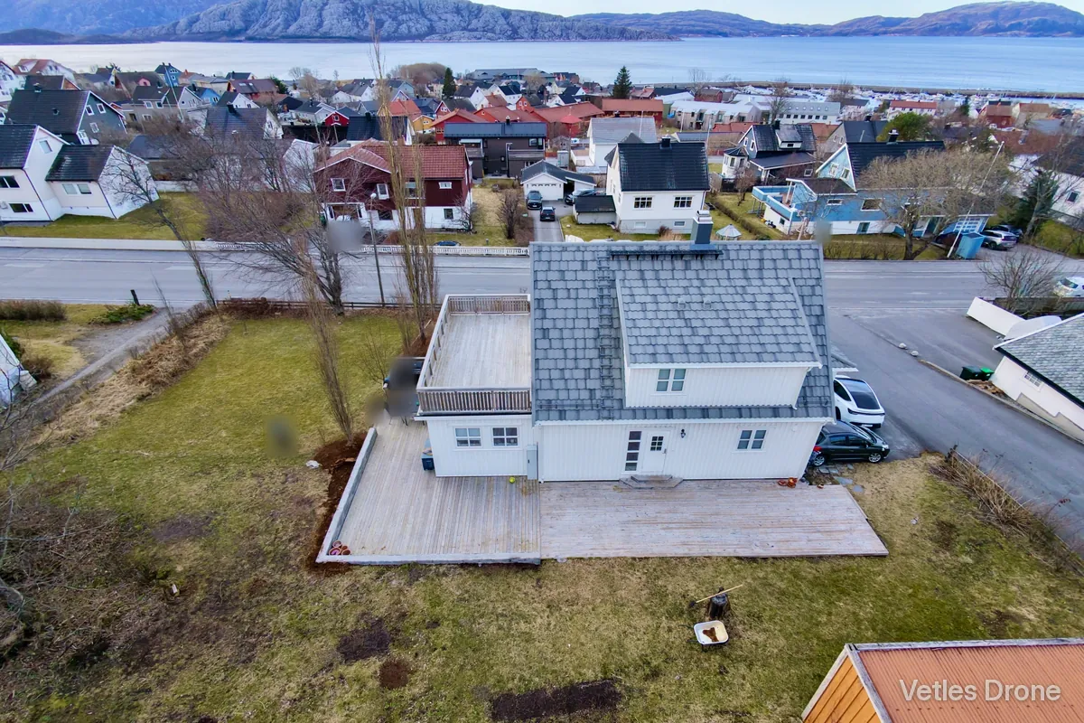 Hvitt hus med grått tak, stor terrasse og hage, fotografert skrått ovenfra med drone, med boligområde og sjø i bakgrunnen