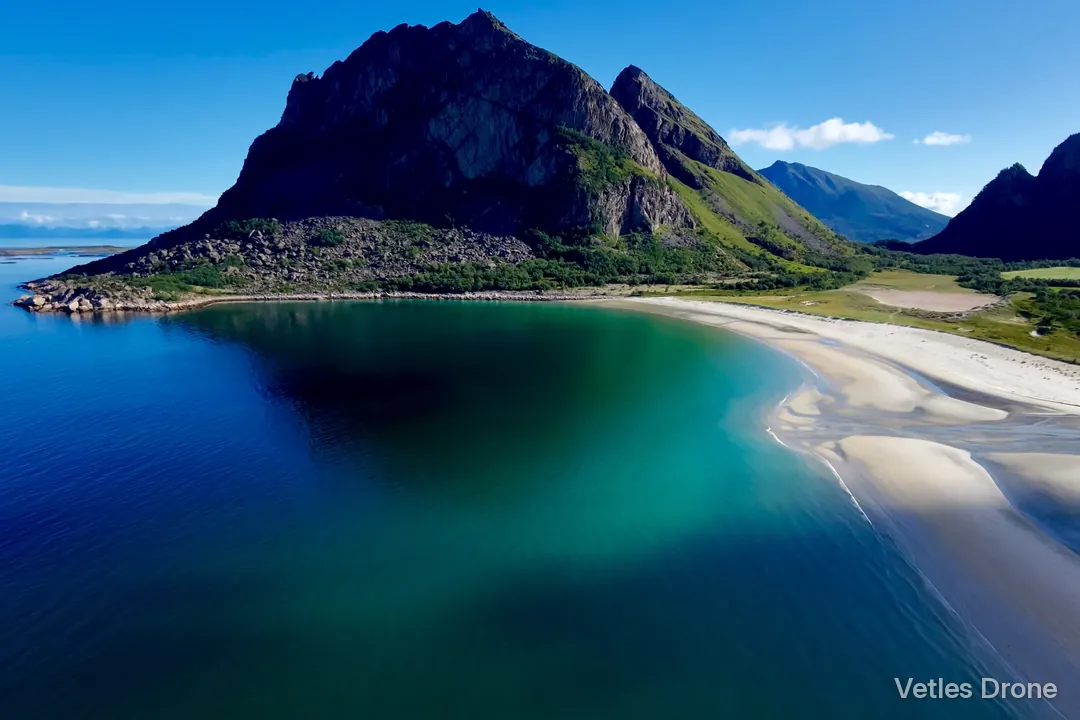 Hvit sandstrand og turkis sjø under spisse fjell en solrik sommerdag, sett fra drone