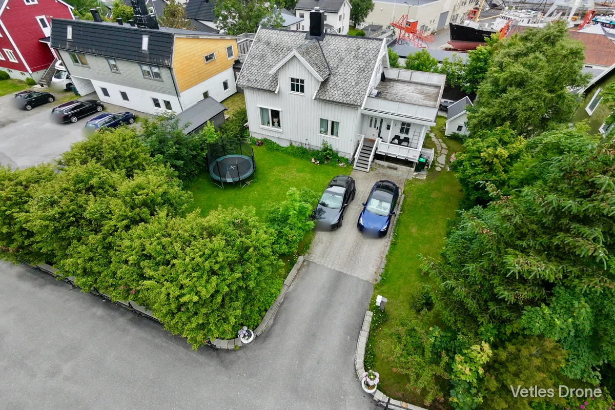 Hvitt hus med skifertak, inngangsparti, innkjørsel og hage, fotografert skrått ovenfra med drone en sommerdag